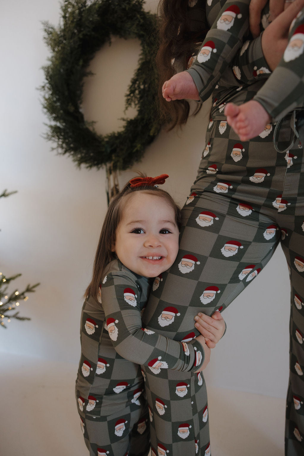 A young child in forever french baby Bamboo Two Piece Pajamas | Santa Checkerboard hugs an adult's leg and smiles, while a baby in matching pajamas is held by the adult. A holiday wreath and decorated tree are in the background.