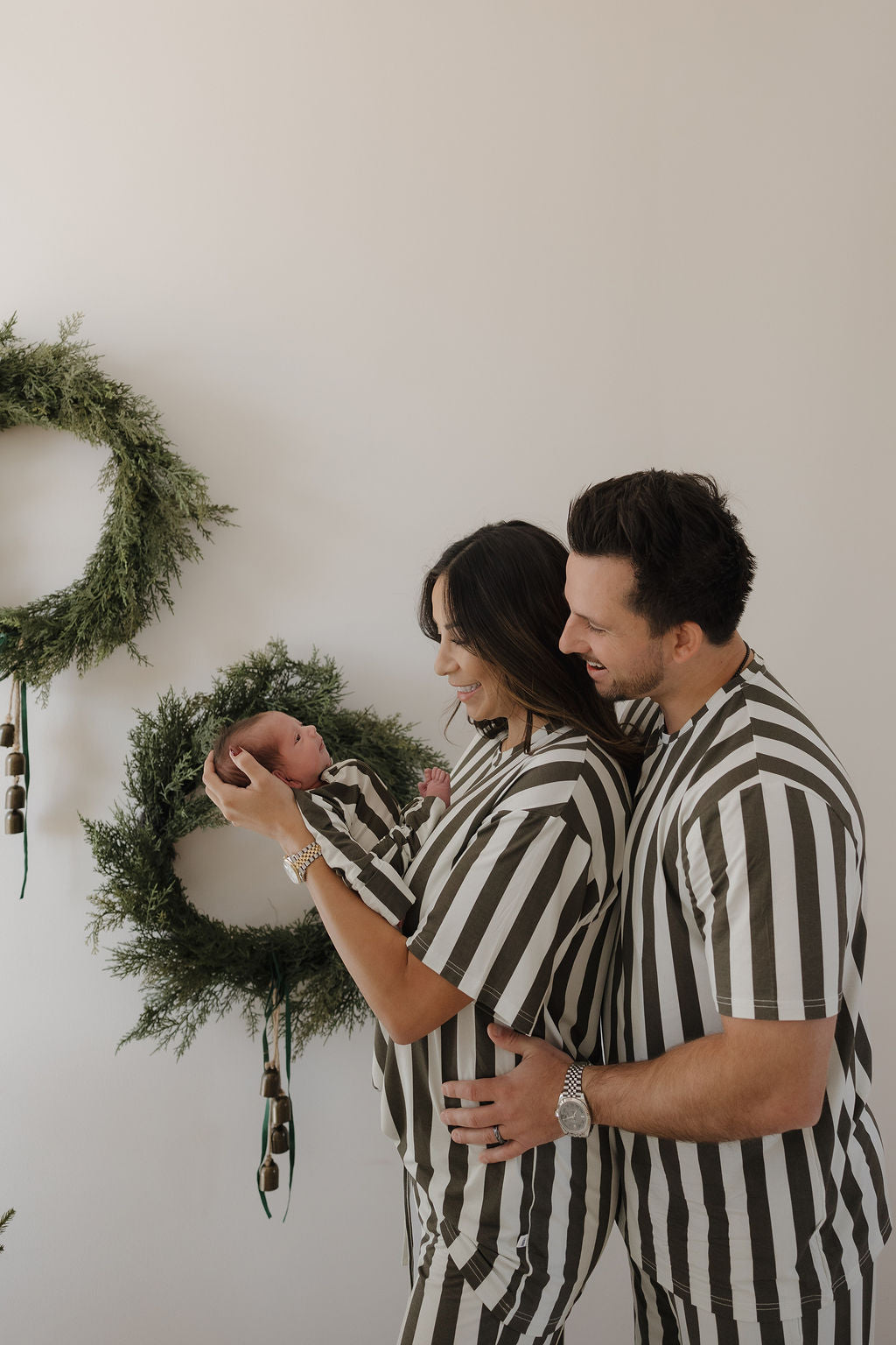 A smiling couple in matching forever french baby Bamboo Zip Pajamas in Fireside Stripe holds their newborn, also dressed in the soft, breathable sleepwear, as they stand before a white wall decorated with green wreaths.