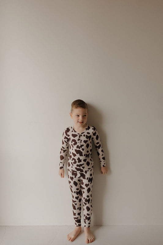 A young boy stands barefoot against a light wall, wearing forever french baby’s Bamboo Two Piece Pajamas | Moo!—long-sleeved with a brown and white cow print—showing off the soft, breathable comfort of bamboo kids sleepwear.