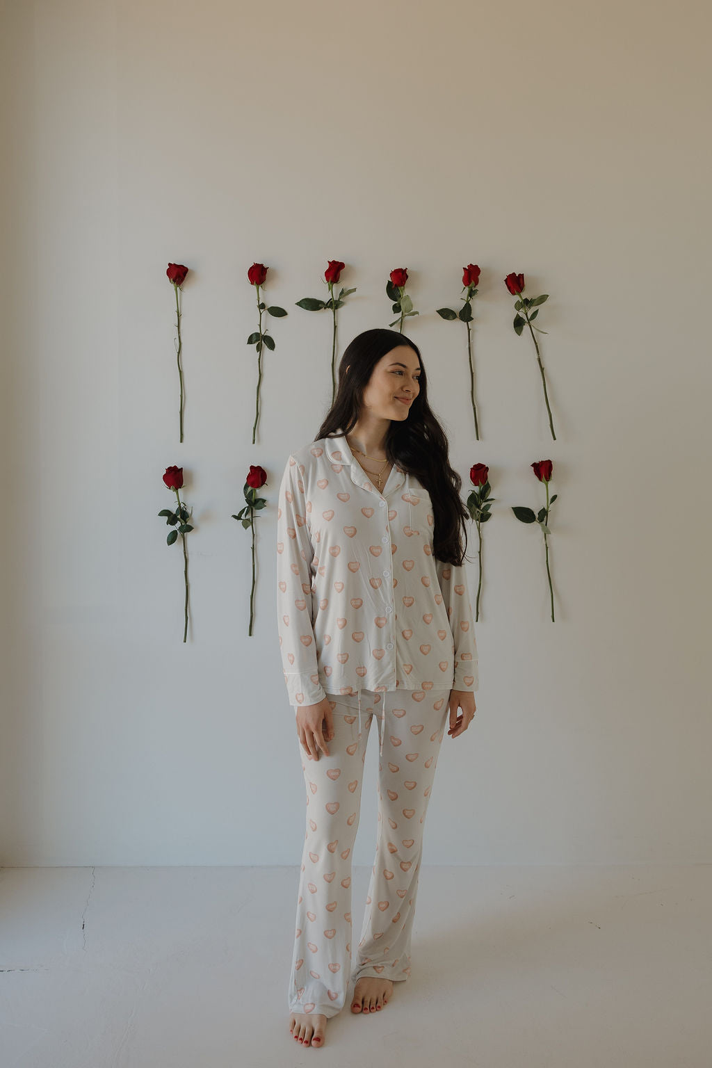 A woman stands barefoot by a white wall with nine red roses, wearing the forever french baby Women's Bamboo Button Up Lounge Set in Love Notes - Grey.