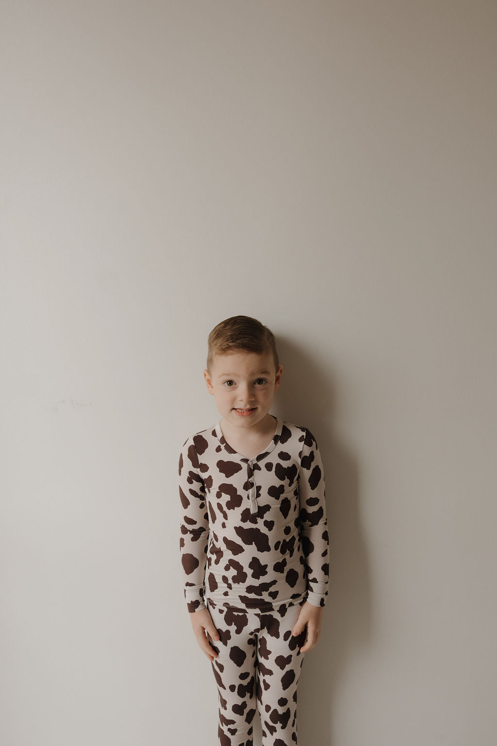 A young boy with short light brown hair smiles softly, wearing forever french baby's Bamboo Two Piece Pajamas | Moo!—matching hypo-allergenic sleepwear in a brown and white cow print, standing against a plain light-colored wall.