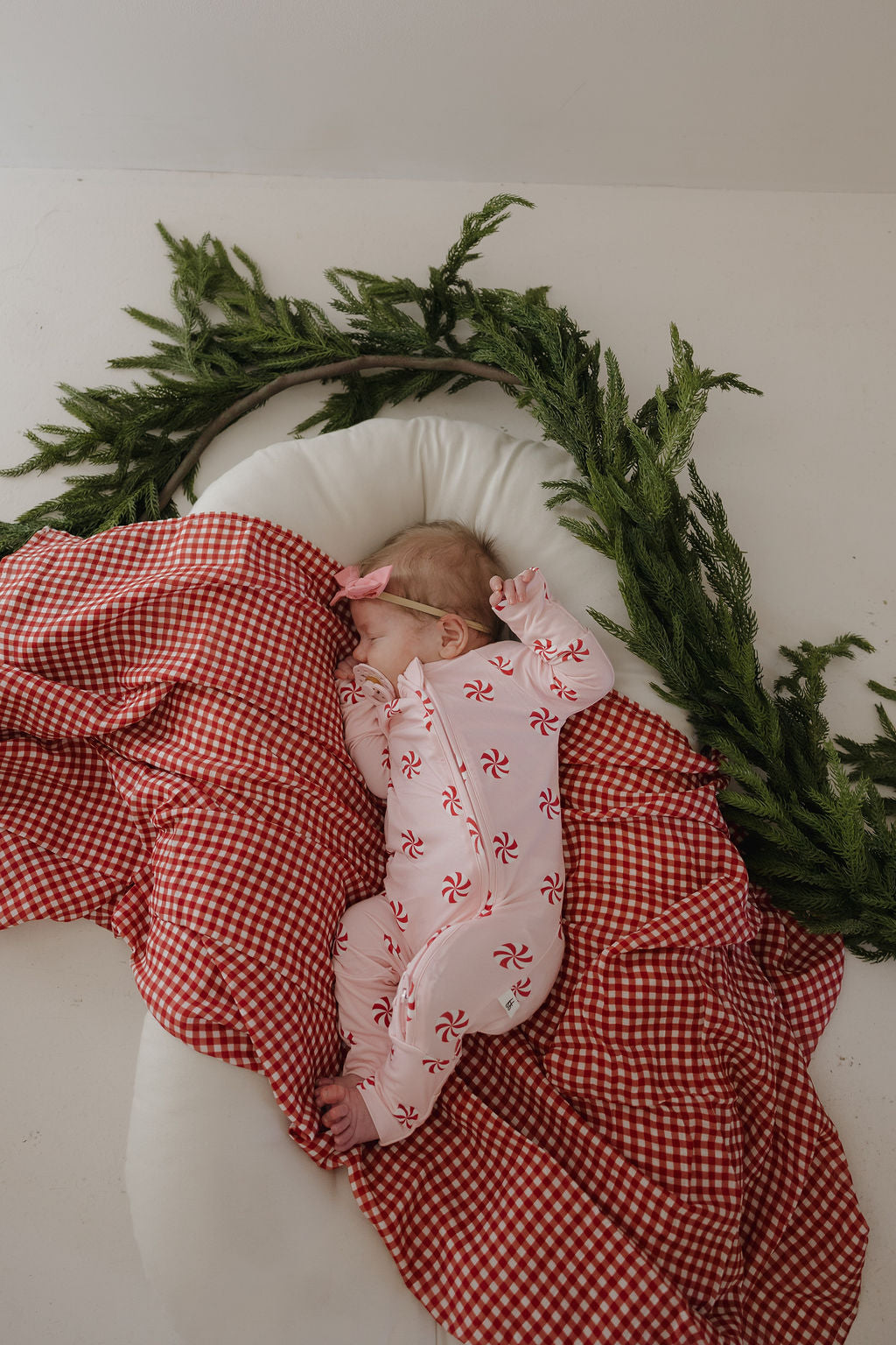 A baby sleeps on a white cushion, partially wrapped in the forever french baby Muslin Swaddle | Red Gingham, wearing a white onesie with red peppermint patterns, with green pine garland arranged in a semi-circle above.