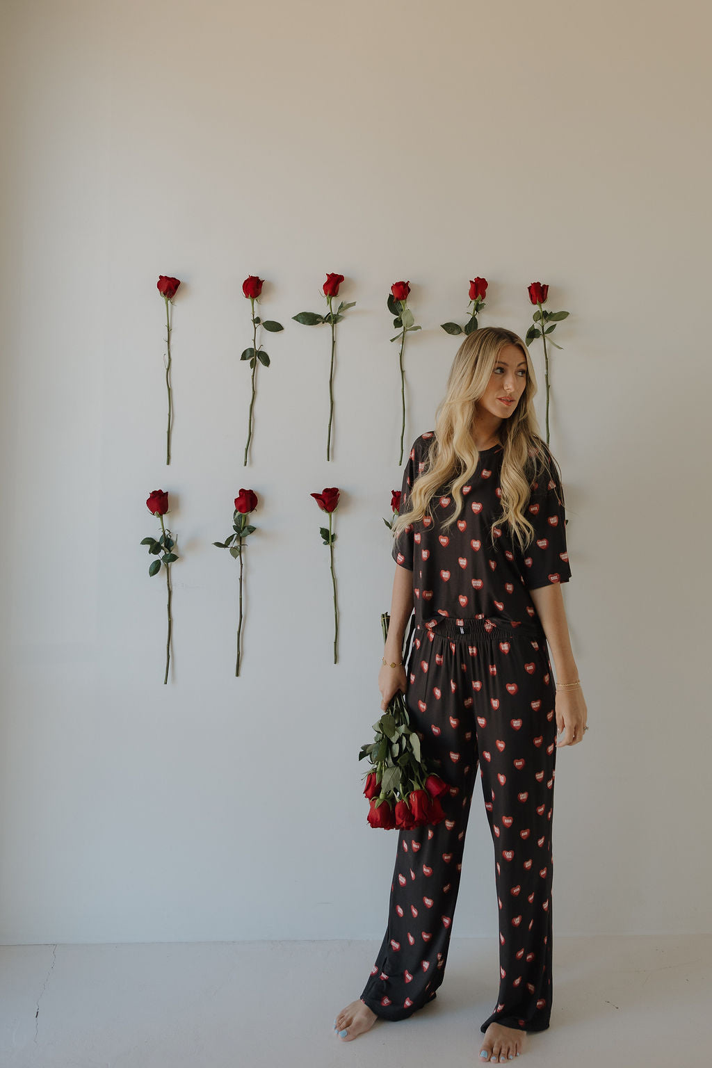 A woman with long blonde hair, barefoot, wears the forever french baby Women's Boxy Tee Lounge Set | Love Notes - Charcoal, featuring red lips. She stands by a plain wall decorated with nine taped red roses and holds a bouquet of red roses.