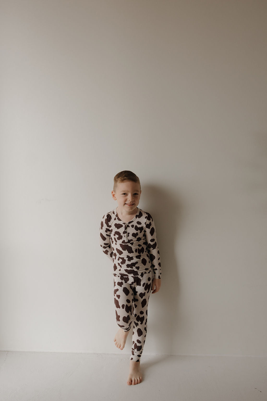 A young boy stands barefoot against a light wall, smiling in matching brown and white Bamboo Two Piece Pajamas | Moo! by forever french baby. His hypo-allergenic kids bamboo sleepwear is comfy, stylish, and perfect for bedtime. One knee is slightly bent.
