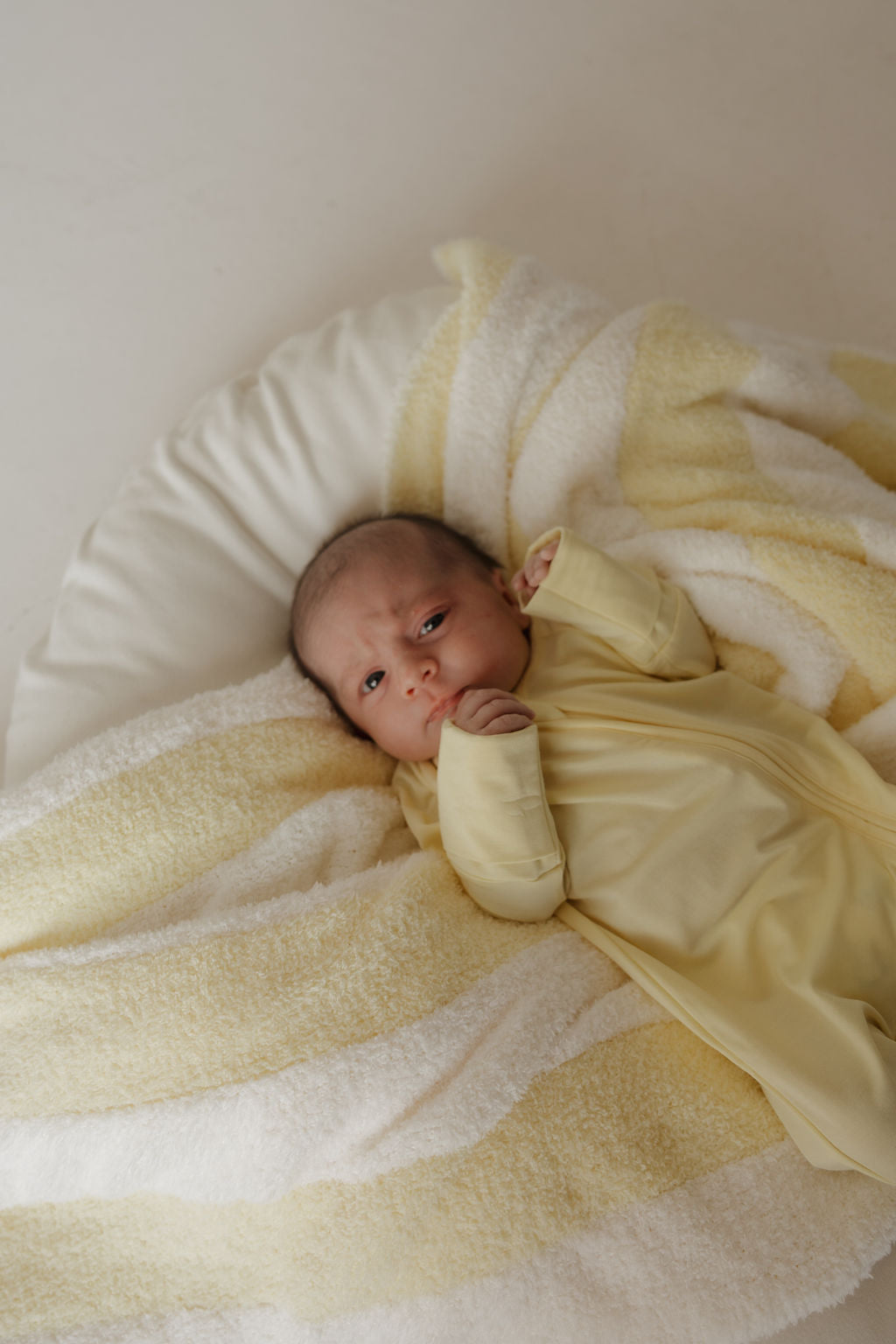 A baby in a yellow outfit lies on a soft, yellow-and-white striped blanket, gazing upward with one hand near their face. The cozy ensemble features the Bamboo Zip Pajamas | Sunny from forever french baby, crafted from breathable bamboo fabric to ensure comfort and gentleness for delicate skin.