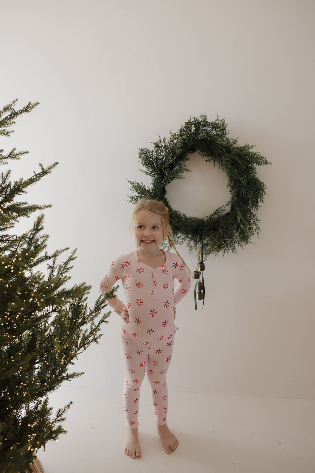A young girl wears forever french baby’s Bamboo Two Piece Pajamas in Twisted Peppermint, smiling with hands on hips in front of a wreath on a white wall. A lit, decorated Christmas tree appears to the left.