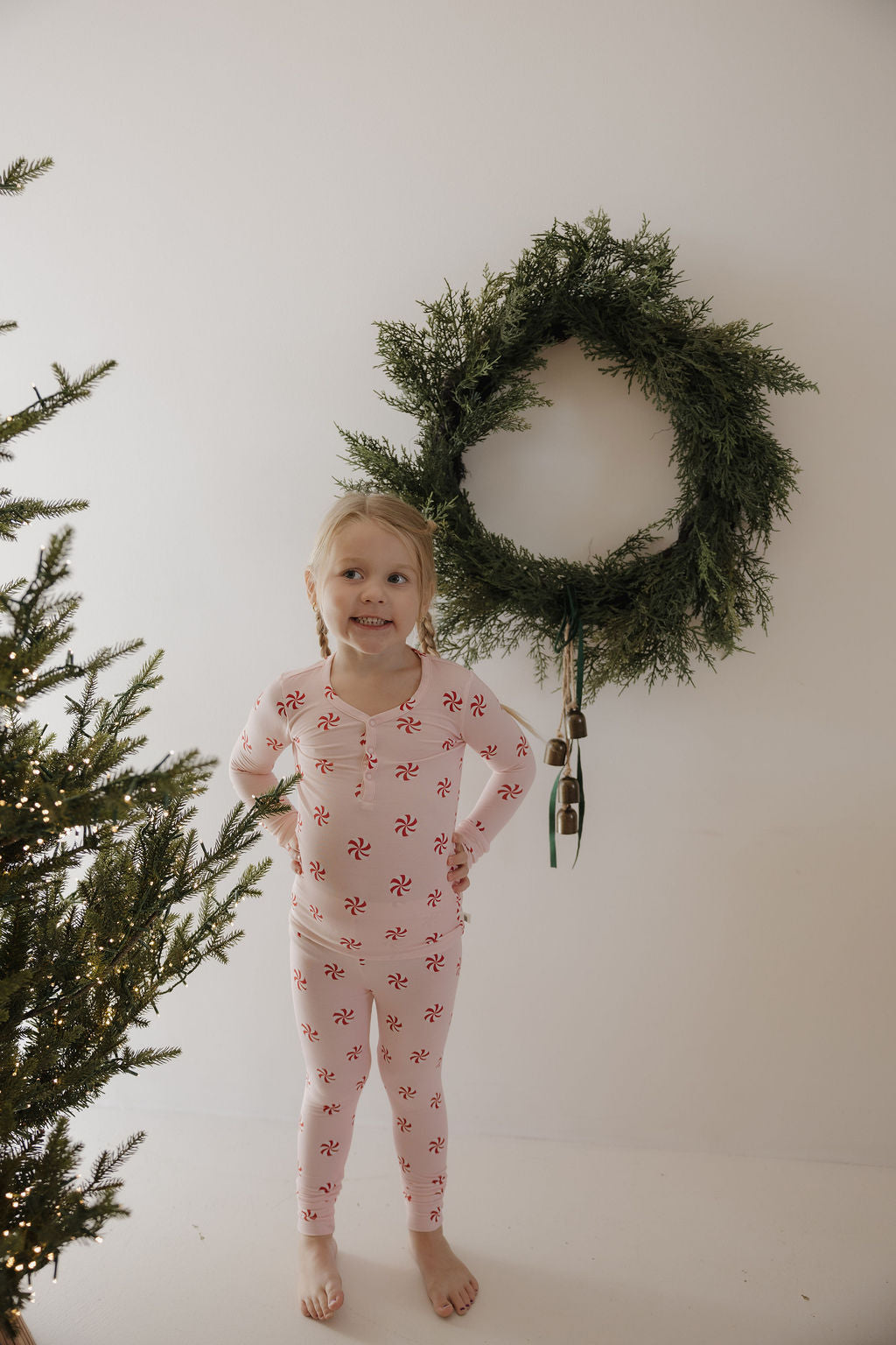 A young child wears forever french baby’s Bamboo Two Piece Pajamas | Twisted Peppermint while standing by a Christmas tree. A green wreath on the white wall behind her highlights this festive, cozy kids sleepwear set.