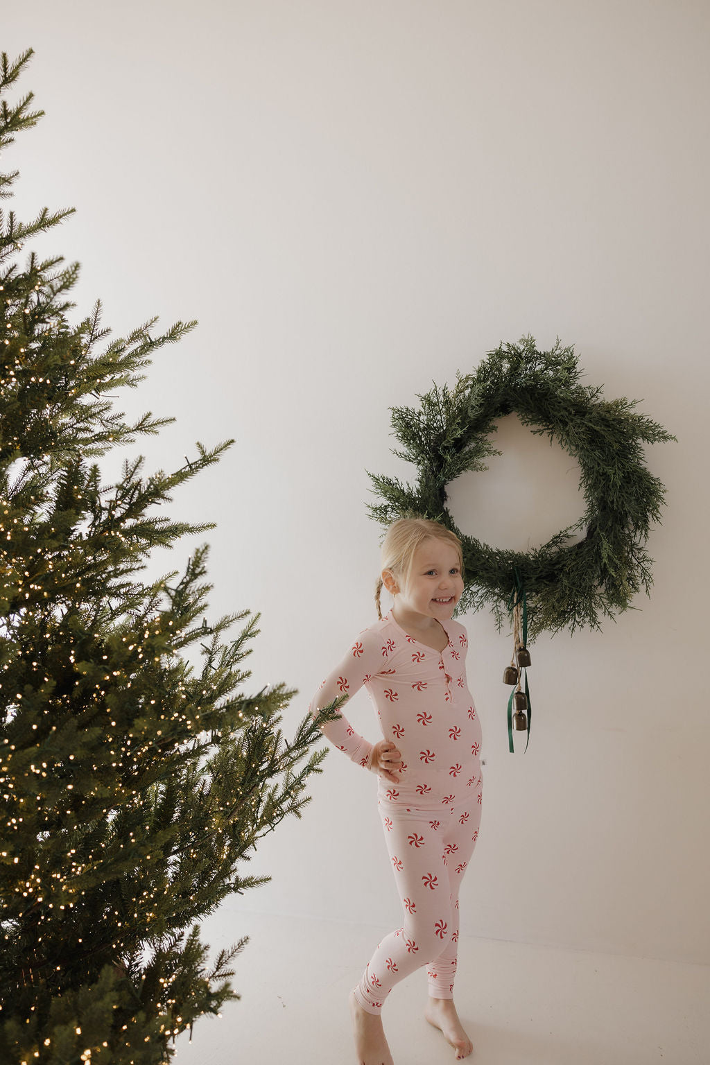 A young child in forever french baby Bamboo Two Piece Pajamas | Twisted Peppermint stands barefoot and smiling near a decorated Christmas tree and green wreath on a white wall, dressed in cozy holiday sleepwear.