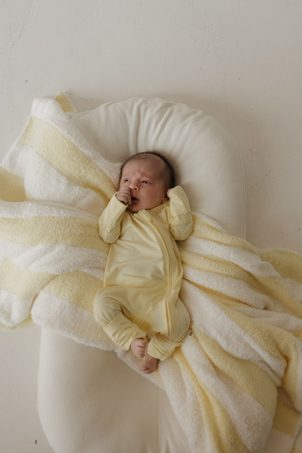A baby in the Bamboo Zip Pajamas | Sunny by forever french baby lies on a white cushion, partially wrapped in a soft yellow and white striped blanket, looking up with one hand near their mouth—highlighting the comfort and gentle touch of these hypoallergenic pajamas.