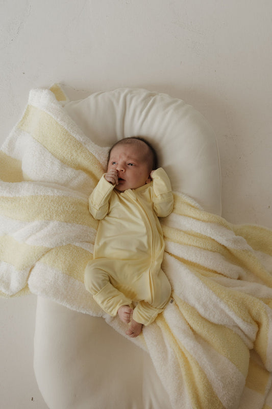 A baby in the Bamboo Zip Pajamas | Sunny by forever french baby lies on a white cushion, partially wrapped in a soft yellow and white striped blanket, looking up with one hand near their mouth—highlighting the comfort and gentle touch of these hypoallergenic pajamas.