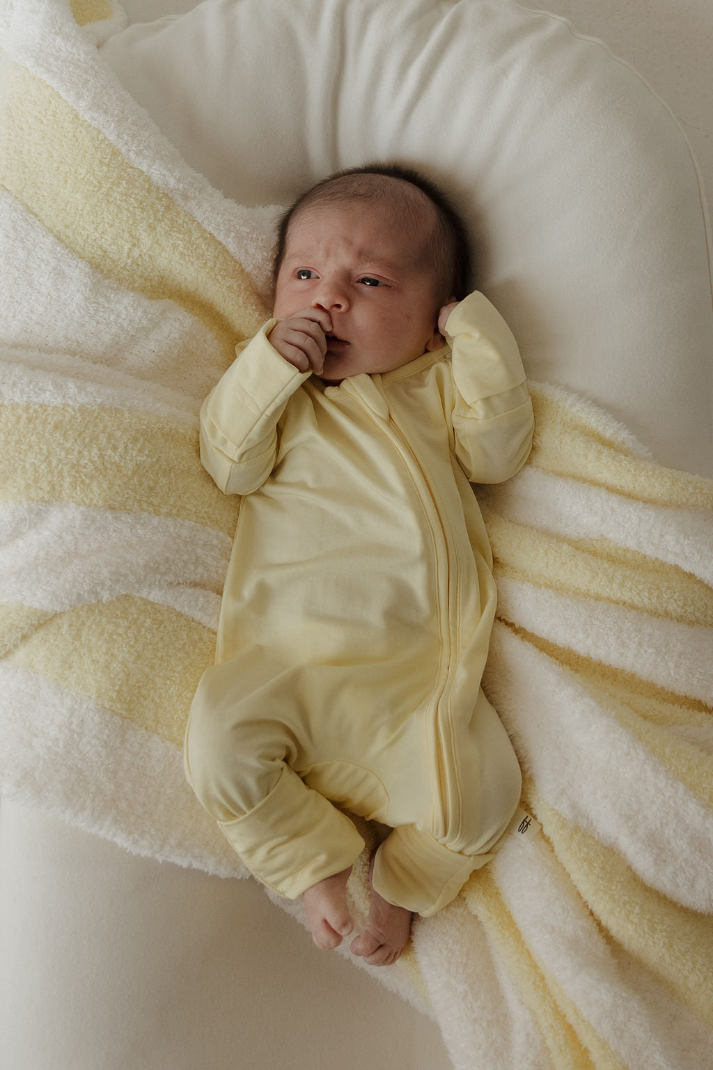 A baby in the forever french baby Bamboo Zip Pajamas | Sunny, a yellow onesie designed for gentle comfort, lies on a soft, cream and yellow-striped blanket, sucking their thumb and looking upward—cozy in hypoallergenic baby clothing.