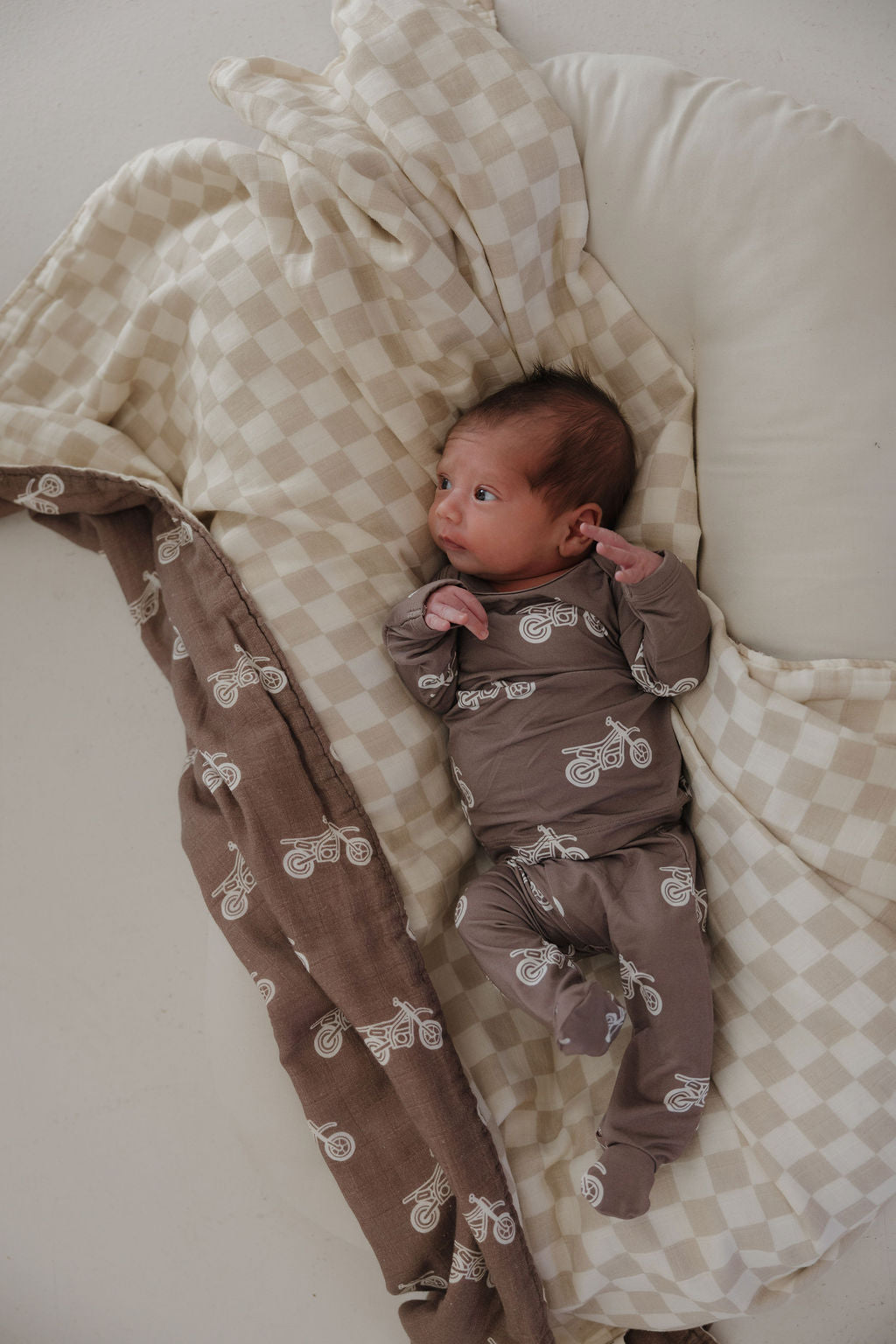 A newborn in the forever french baby Sleepy Time Set | Dirt Bike—a brown, hypoallergenic outfit with white bike prints—lies on a cream and beige checkered blanket, gazing up with wide eyes.