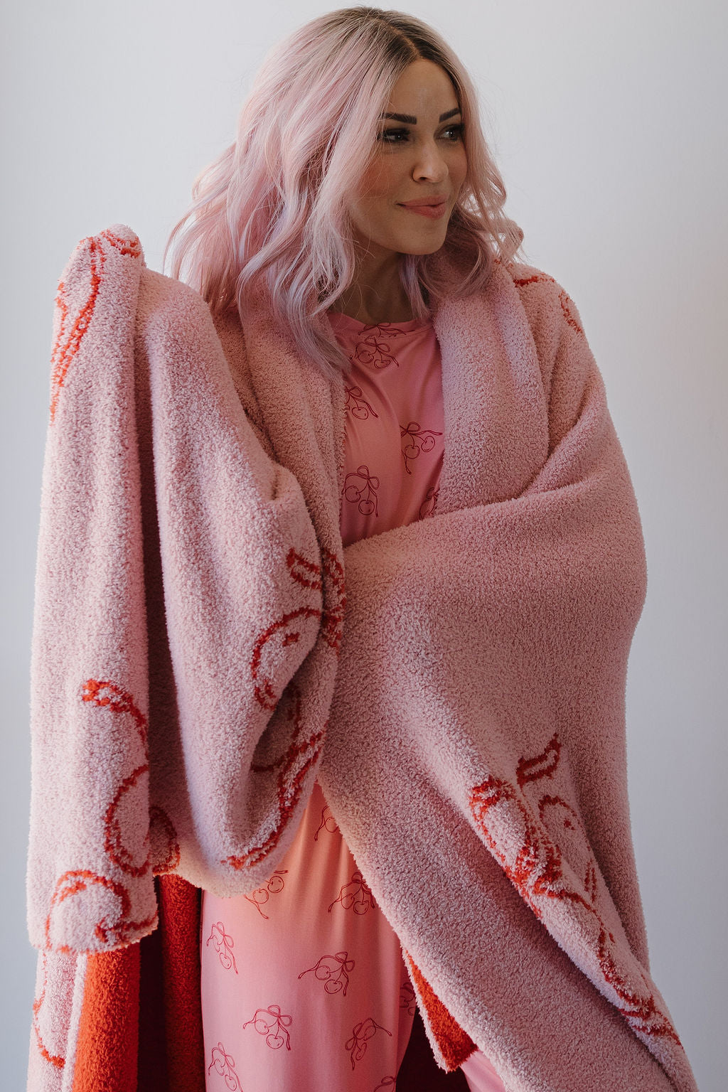 A woman with long, wavy pastel pink hair in a pink patterned dress wraps herself in the Minty Cherries Plush Blanket | Iconic Cherry, an Oeko-Tex certified microfiber blanket with red cherry designs, smiling softly against a plain white background.