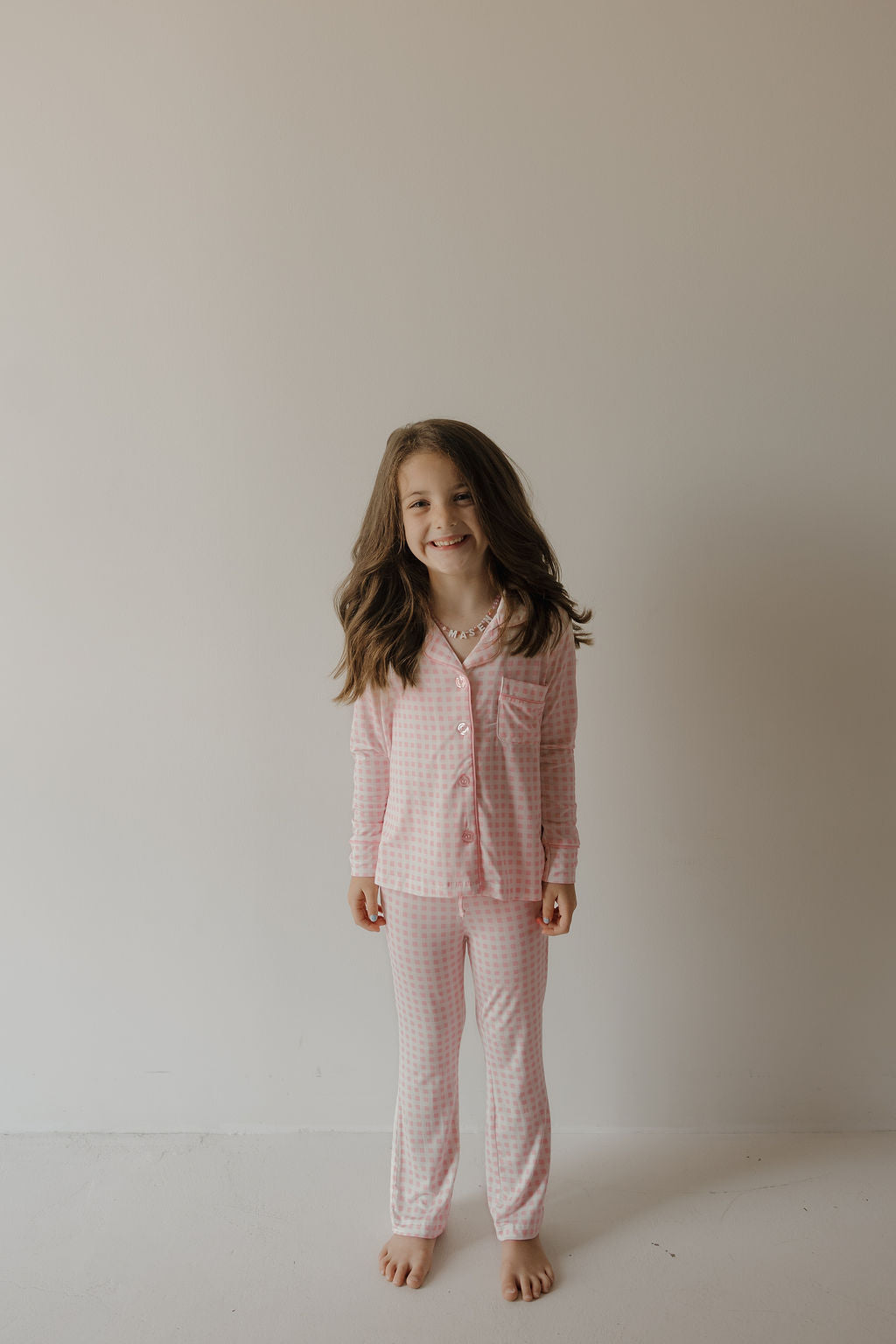 A young girl with long brown hair smiles while standing barefoot against a plain white wall, wearing the Girl’s Bamboo Button Up Lounge Set | Giselle by forever french baby, crafted from soft, hypo-allergenic bamboo fabric.