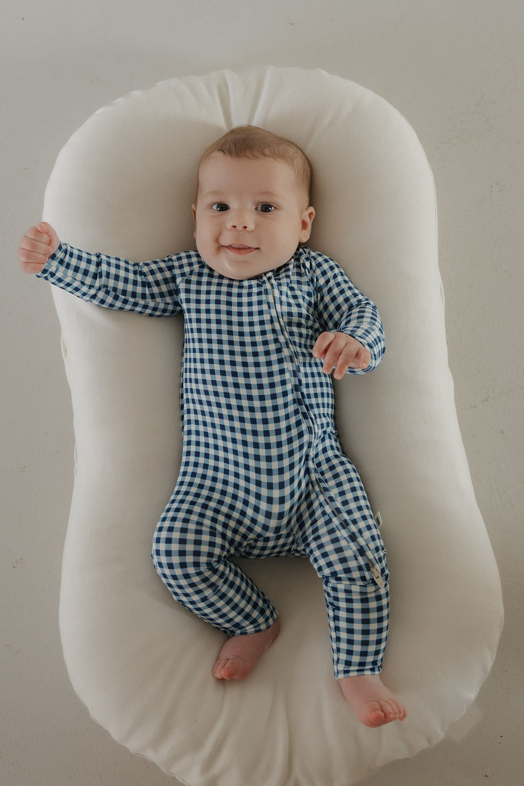 A baby in forever french baby's Bamboo Zip Pajamas in Blue Gingham lies on a soft white cushion, smiling and looking up with one arm outstretched in this cozy, hypoallergenic onesie.