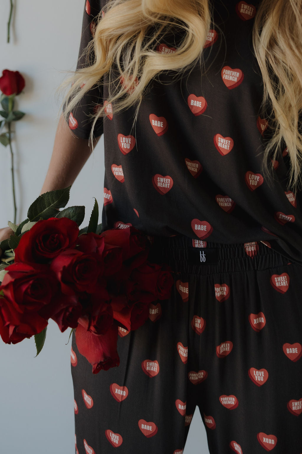 A woman with long blonde hair holds red roses while wearing the forever french baby Women's Boxy Tee Lounge Set | Love Notes - Charcoal, featuring red hearts and romantic words. More roses appear in the background.