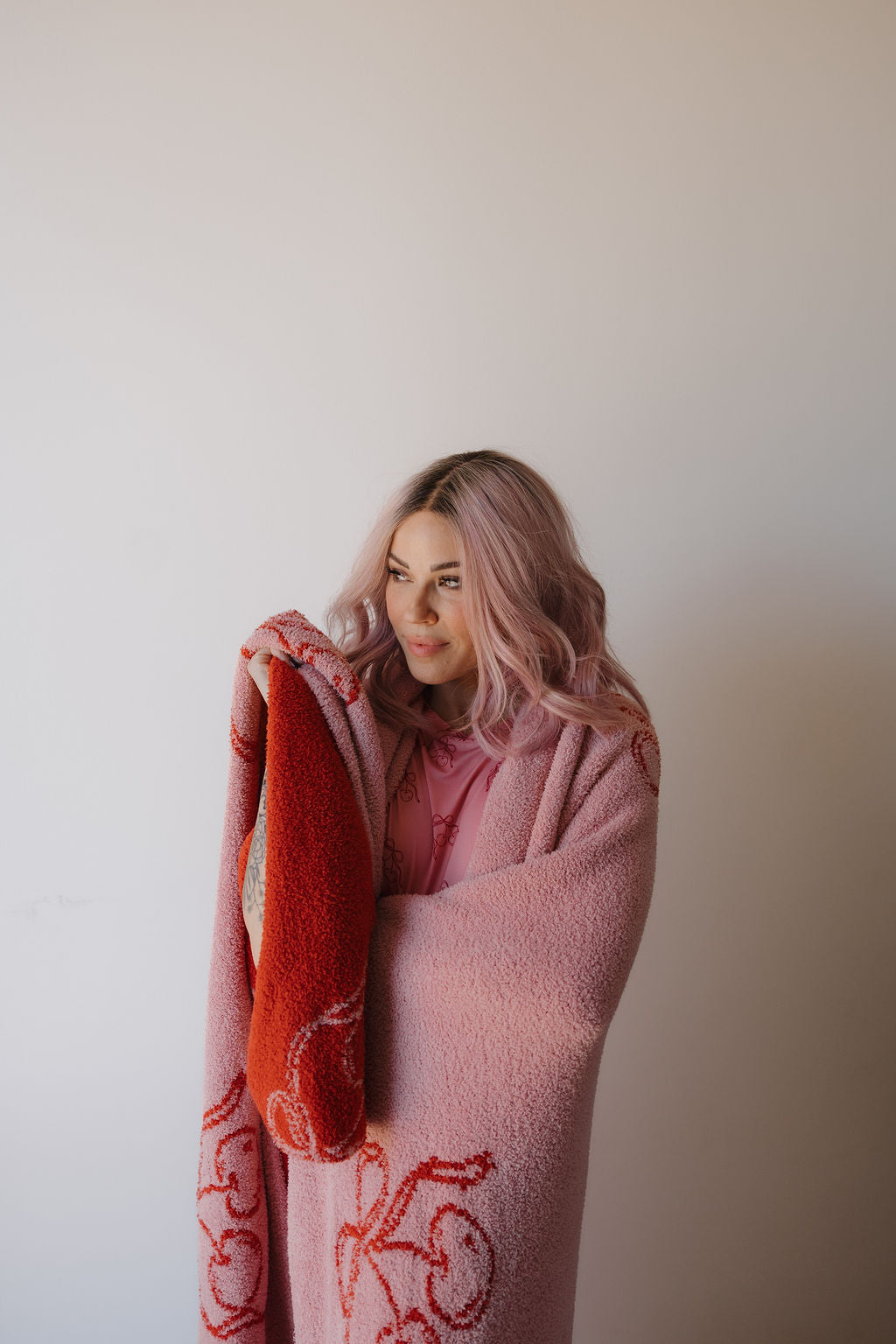 A woman with long, wavy pink hair stands against a plain light wall, wrapped in the Minty Cherries Plush Blanket | Iconic Cherry—an Oeko-Tex certified pink and red throw with a subtle pattern. She gazes off to the side with a relaxed expression.