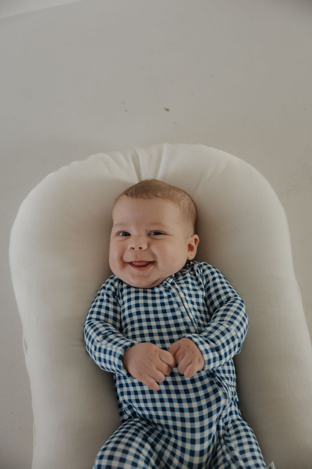 A smiling baby in forever french baby Bamboo Zip Pajamas | Blue Gingham lies on a soft white cushion, looking up at the camera.