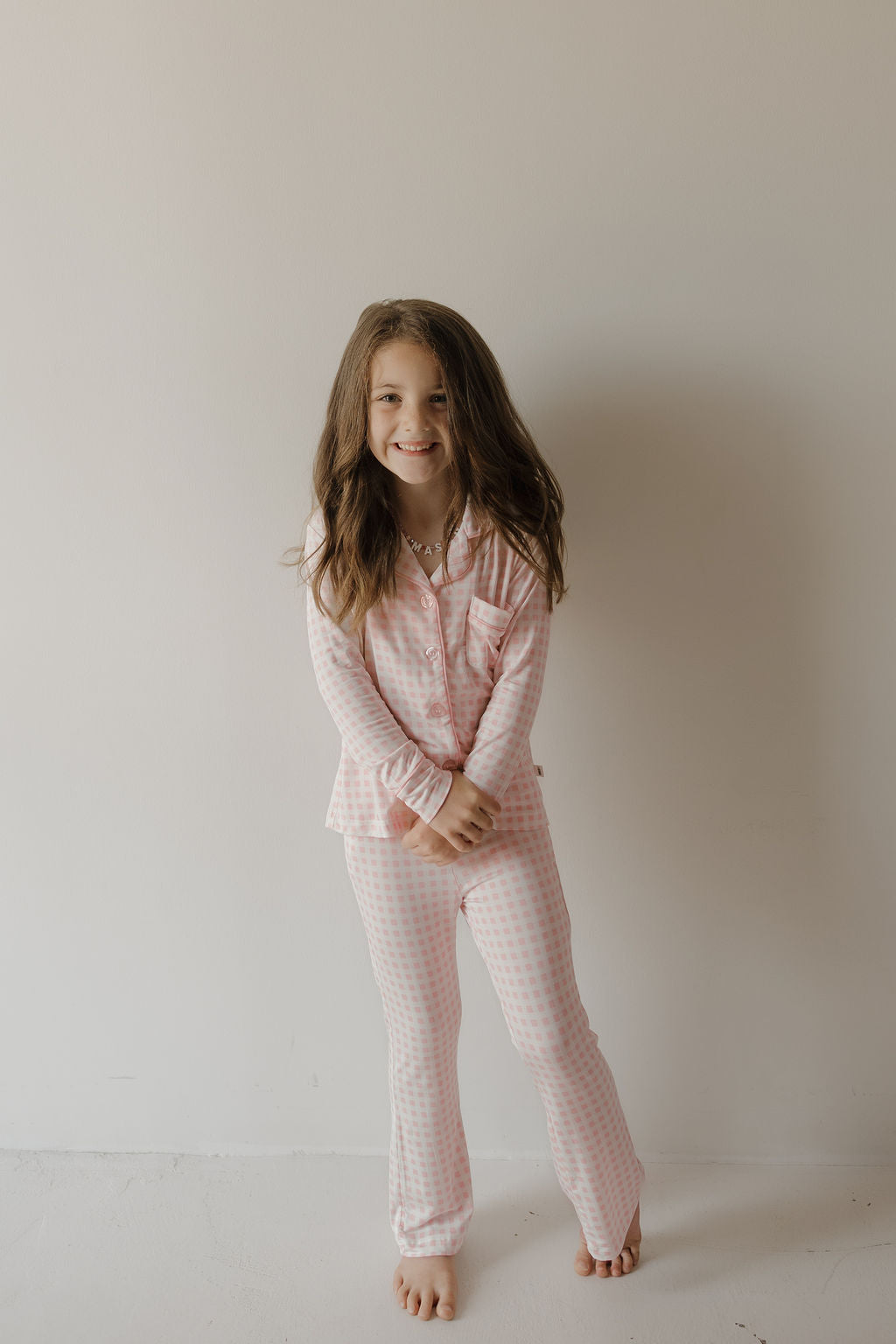 A young girl with long brown hair smiles as she stands barefoot against a plain light wall, hands clasped in front of her. She is wearing the forever french baby Girl’s Bamboo Button Up Lounge Set | Giselle, a matching pink and white checkered toddler set known for its hypo-allergenic bamboo fabric.