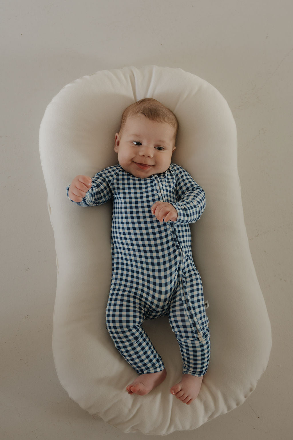 A baby wearing forever french baby Bamboo Zip Pajamas in Blue Gingham smiles while lying on a soft white cushion, looking up.