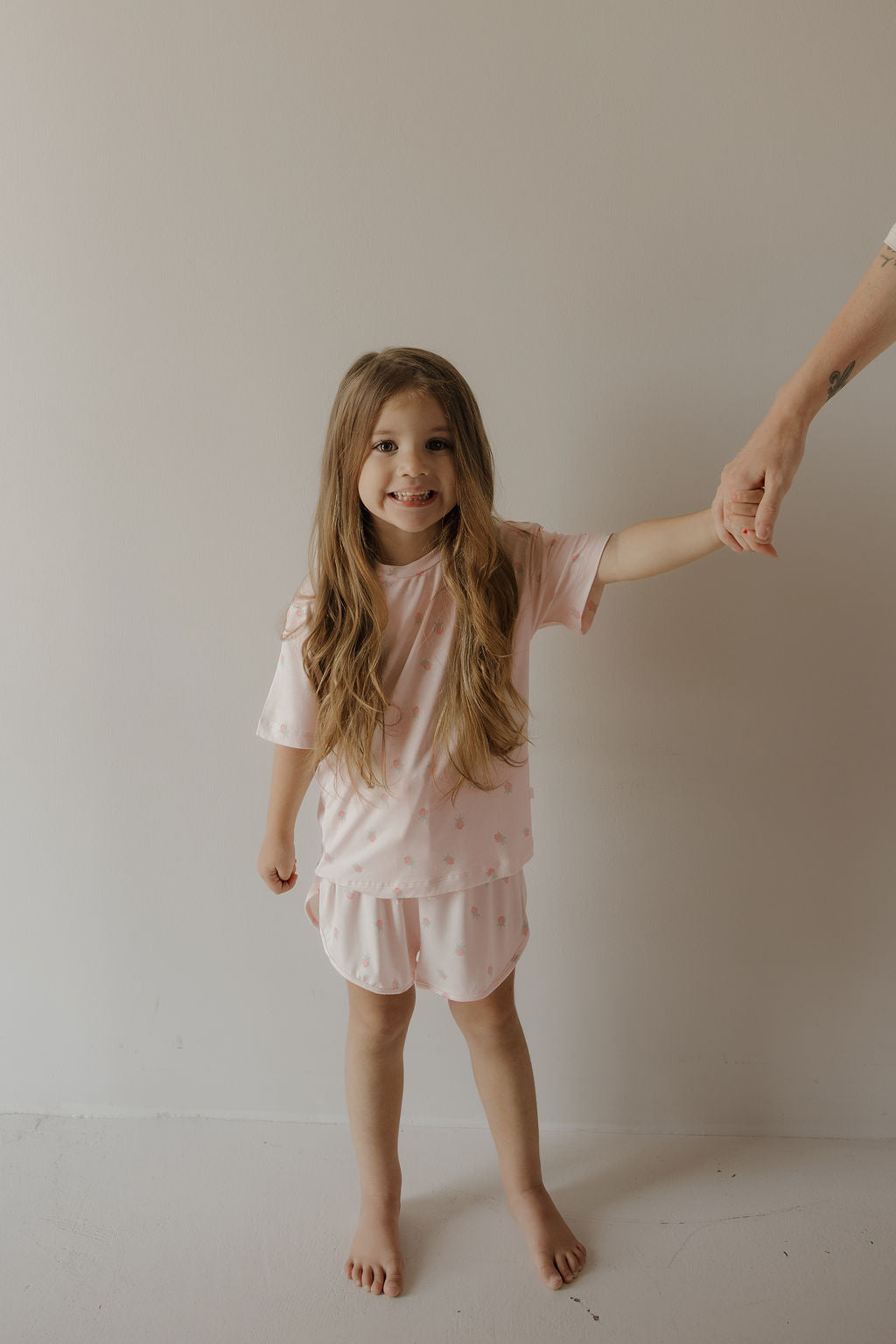 A young girl with long hair, barefoot and smiling, stands against a plain light-colored wall while holding an adult’s hand, wearing the forever french baby Girl’s Bamboo Short Set | Rosie—a hypo-allergenic, light pink outfit.