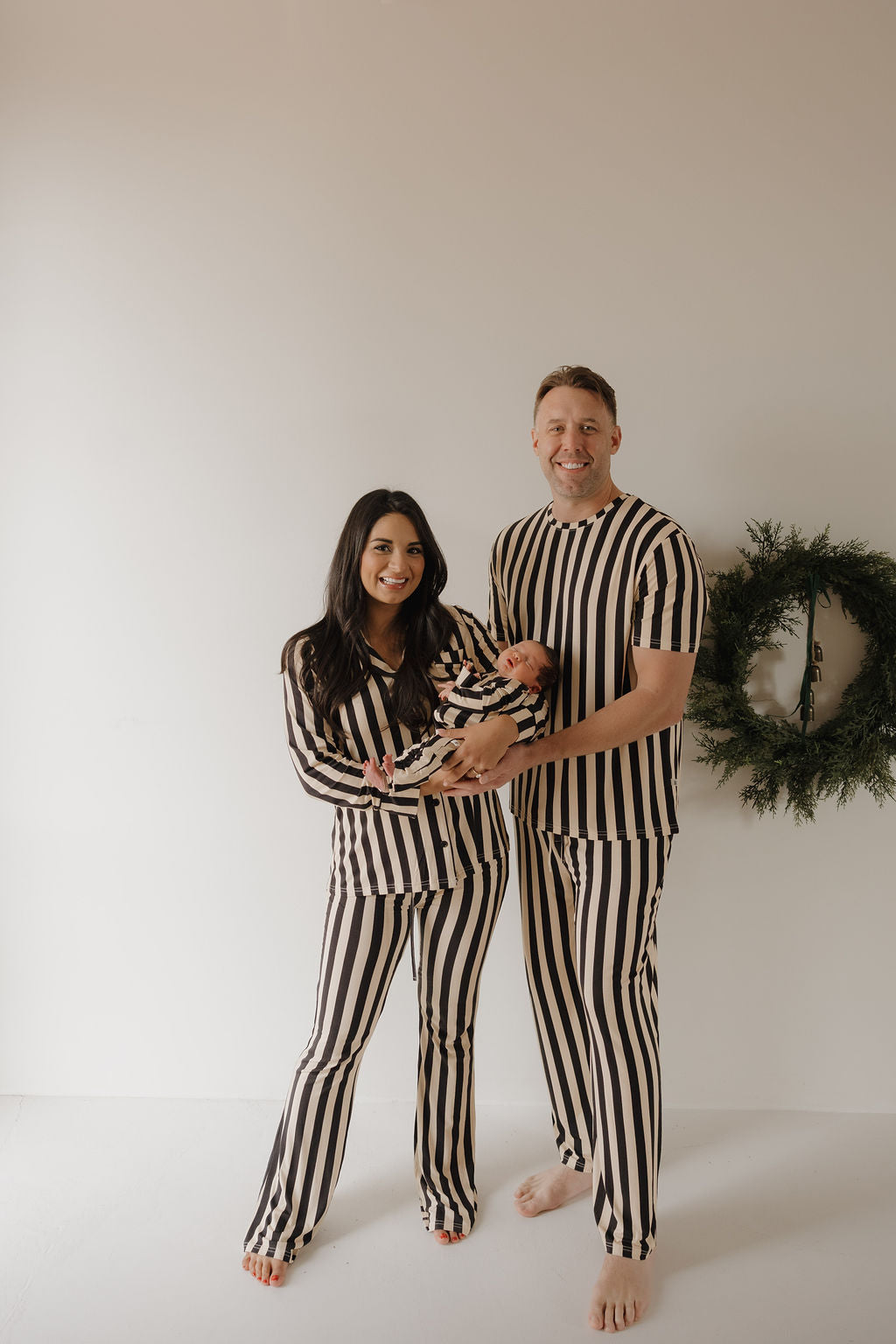 A smiling couple in matching forever french baby Bamboo Zip Pajamas | Midnight Stripe stands barefoot in a minimal room, holding a baby in breathable sleepwear. A green wreath hangs on the wall behind them.