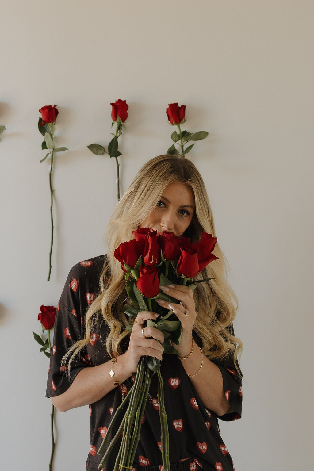 A woman with long blonde hair smiles gently, holding a bouquet of red roses in front of a white wall decorated with single red roses. She wears the Women's Boxy Tee Lounge Set | Love Notes - Charcoal by forever french baby.