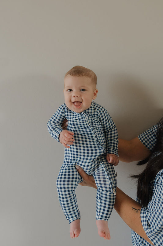 A smiling barefoot baby in forever french baby's Bamboo Zip Pajamas in Blue Gingham is held by an adult, arm visible, facing the camera against a plain light-colored background.