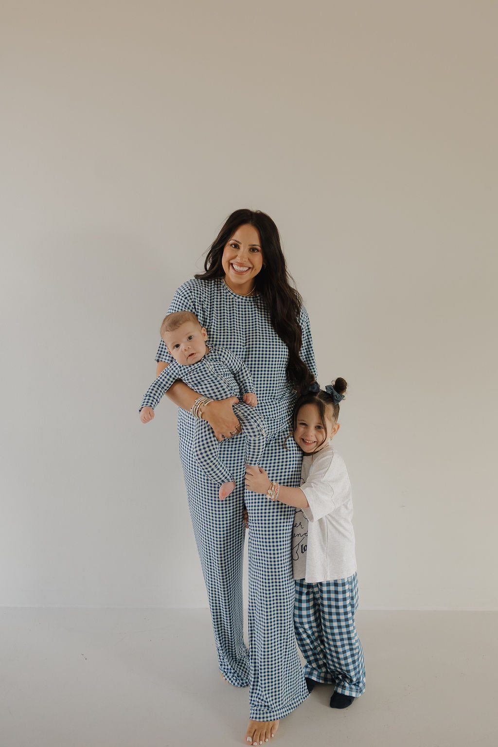 A smiling woman holds a baby, both in forever french baby Bamboo Zip Pajamas in Blue Gingham. A young child, wearing matching blue gingham pants and a white shirt, hugs her leg against a plain light background.