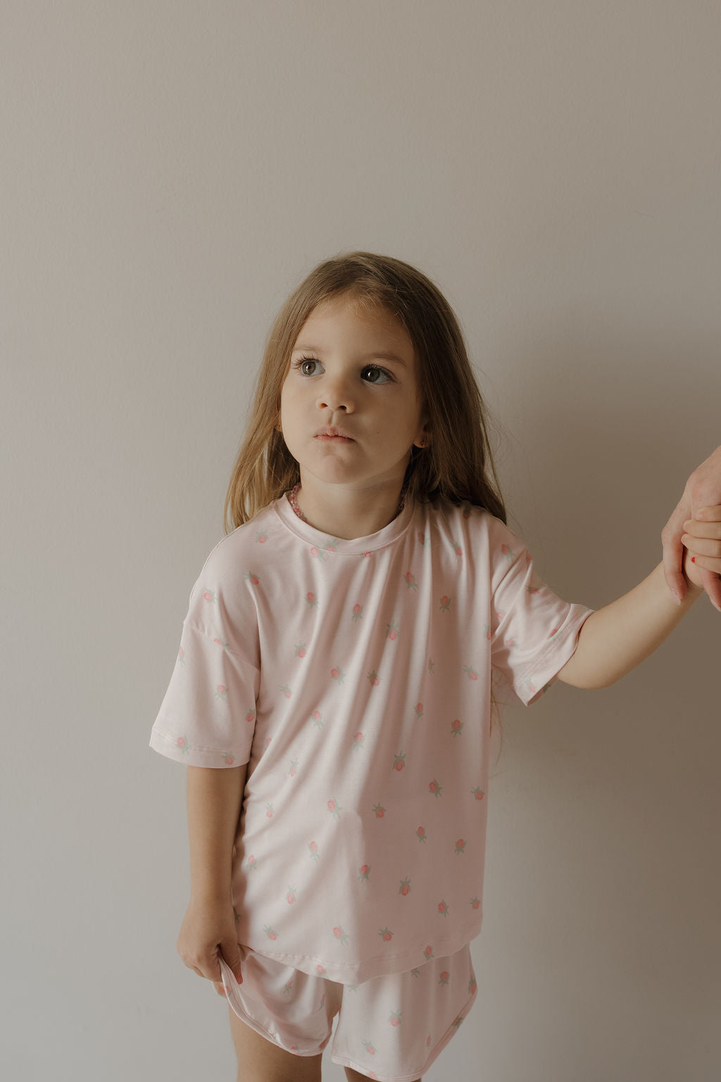 A young girl with long brown hair, wearing the forever french baby Girl’s Bamboo Short Set in Rosie—a perfect example of hypo-allergenic clothing—stands against a plain light-colored wall while holding an adult’s hand.