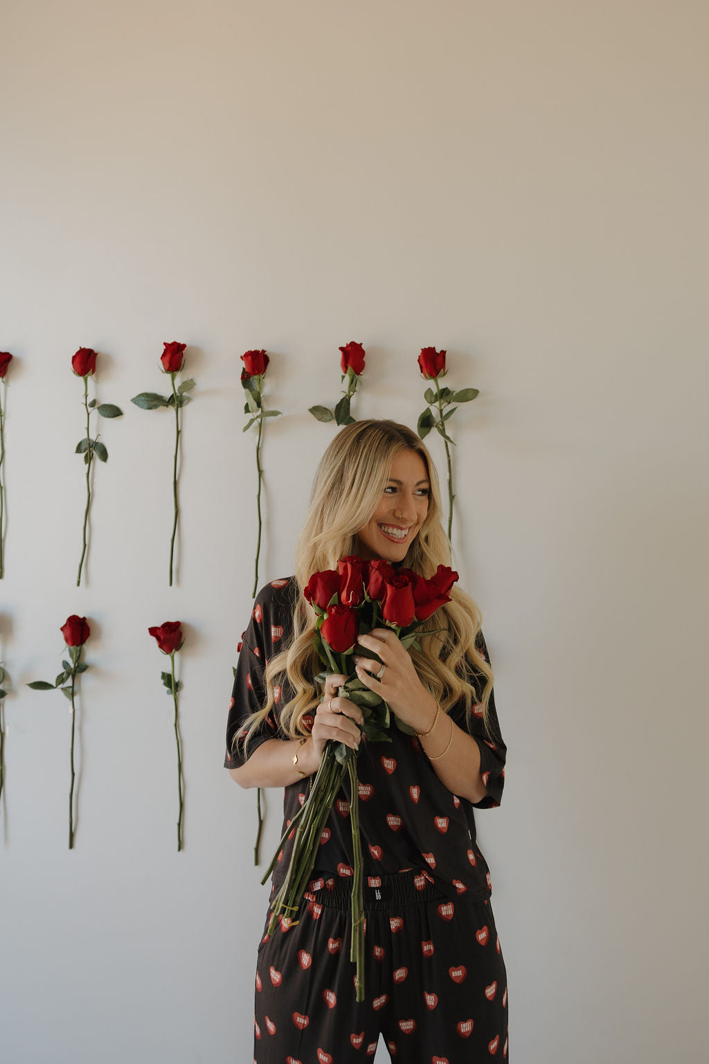 A woman with long blonde hair, wearing the forever french baby Women's Boxy Tee Lounge Set | Love Notes - Charcoal with red heart patterns, smiles holding red roses. Rows of single red roses are taped to a plain white wall behind her.