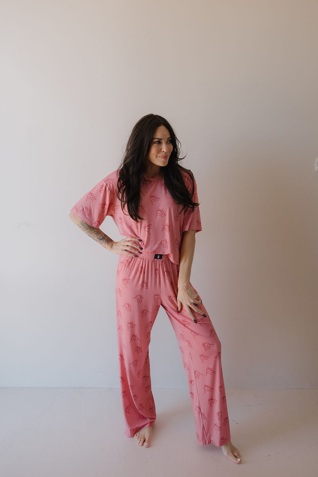 A woman with long dark hair stands barefoot against a light wall, wearing the Minty Cherries Women's Boxy Tee Lounge Set in Iconic Cherry. She has one hand on her hip and smiles slightly.