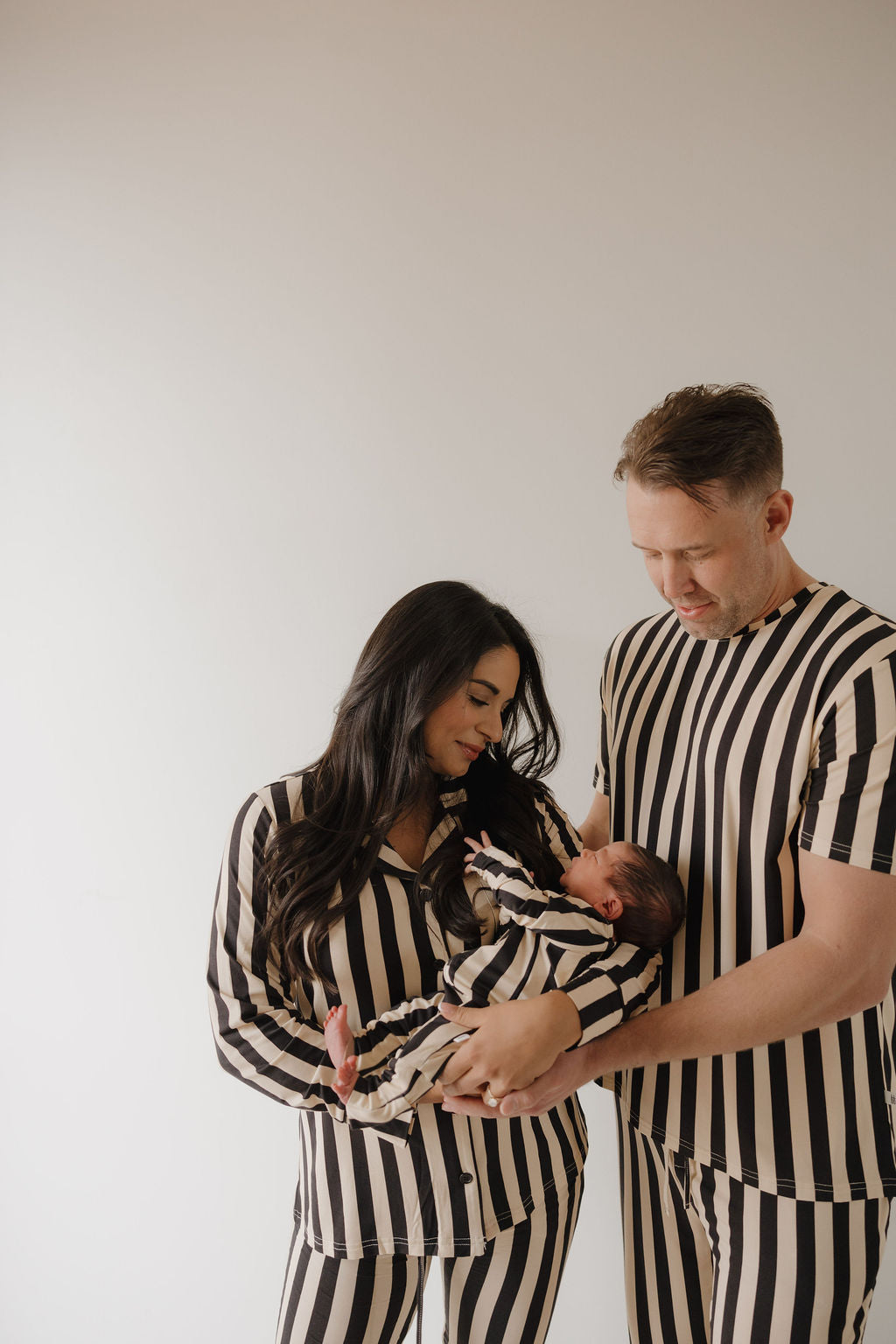 A couple stands together wearing forever french baby’s Midnight Stripe Men’s Short Sleeve Pajamas, lovingly holding a baby dressed in matching hypoallergenic sleepwear against a simple light background.
