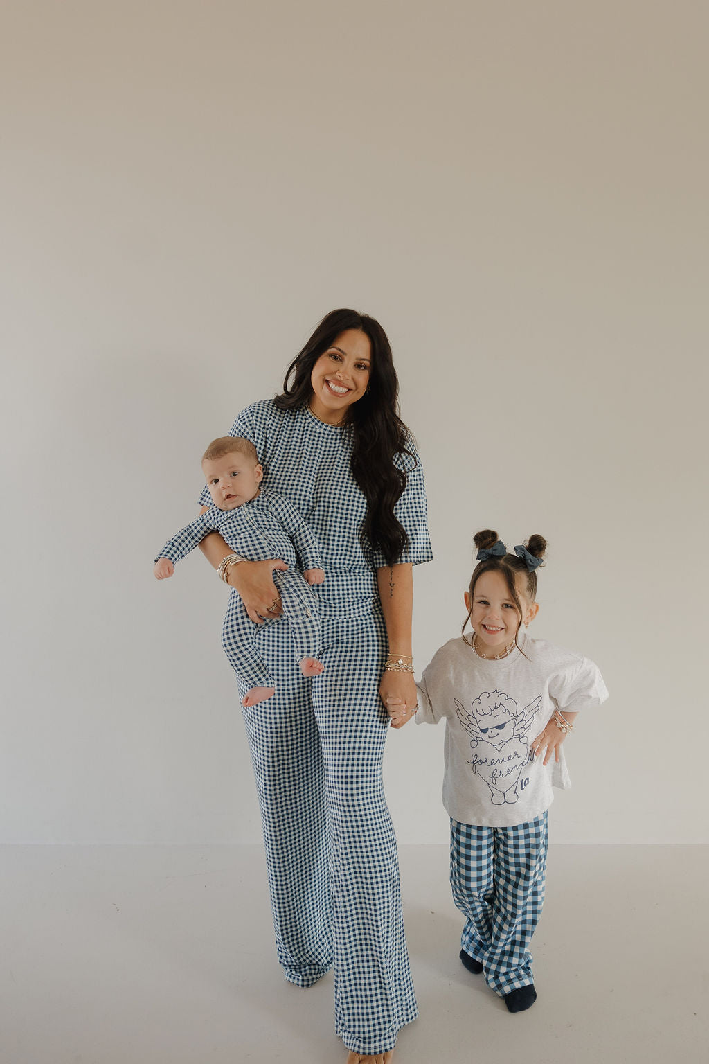 A woman smiles, holding a baby in forever french baby’s Bamboo Zip Pajamas in Blue Gingham, standing beside a young girl in matching checkered pants and a white shirt. These soft, breathable pajamas keep little ones cozy and stylish.
