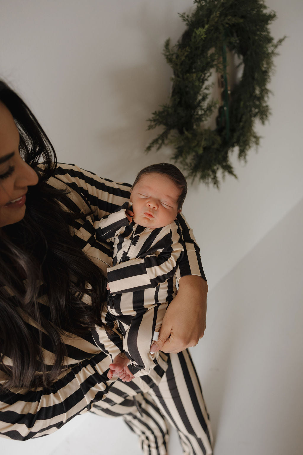 A woman holds a sleeping baby, both dressed in forever french baby Midnight Stripe bamboo sets, with a green wreath on the white wall behind them.