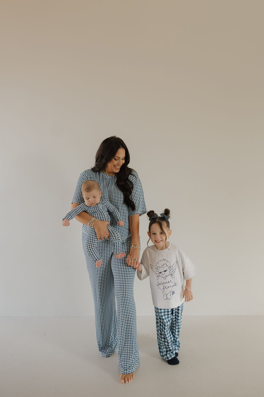 A barefoot woman holds a baby in forever french baby Bamboo Zip Pajamas | Blue Gingham and clasps hands with a smiling young girl, both in matching checkered pajamas against a plain beige background.