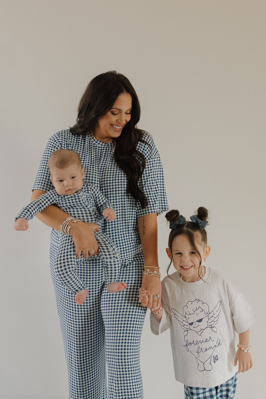 A woman in the forever french baby Women's Boxy Tee Lounge Set in Blue Gingham holds a baby in matching attire and smiles at a girl wearing a white "forever friend" angel tee. They are posed against a plain, light background.