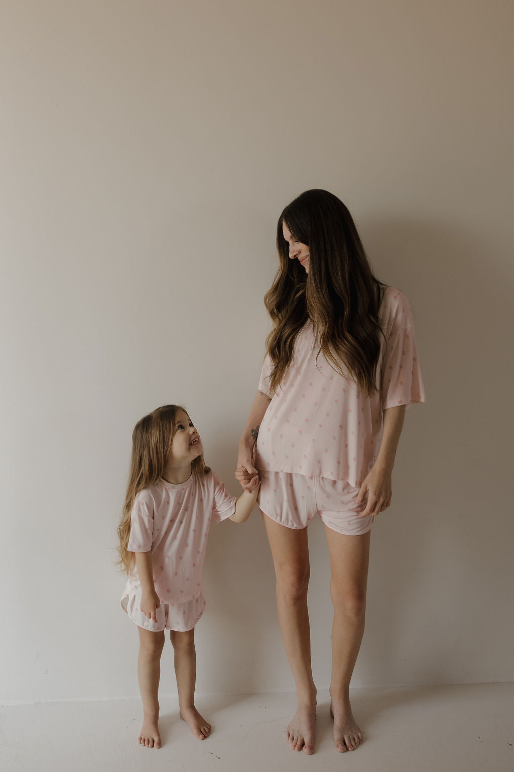 A woman and a young girl, both barefoot and smiling warmly as they stand against a plain wall, wear matching pink polka dot pajamas made from soft, hypo-allergenic bamboo fabric. The young girl looks up at the woman and holds her hand while wearing the Girl’s Bamboo Short Set | Rosie by forever french baby.