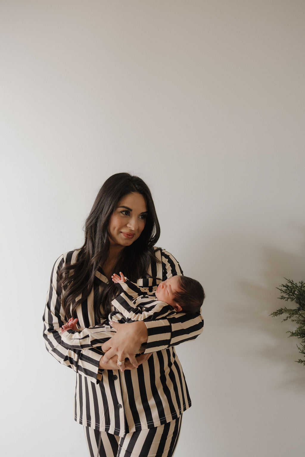 A woman with long dark hair wears the forever french baby Women's Bamboo Button Up Lounge Set in Midnight Stripe, holding a newborn dressed in matching hypo-allergenic bamboo clothing against a plain light background.
