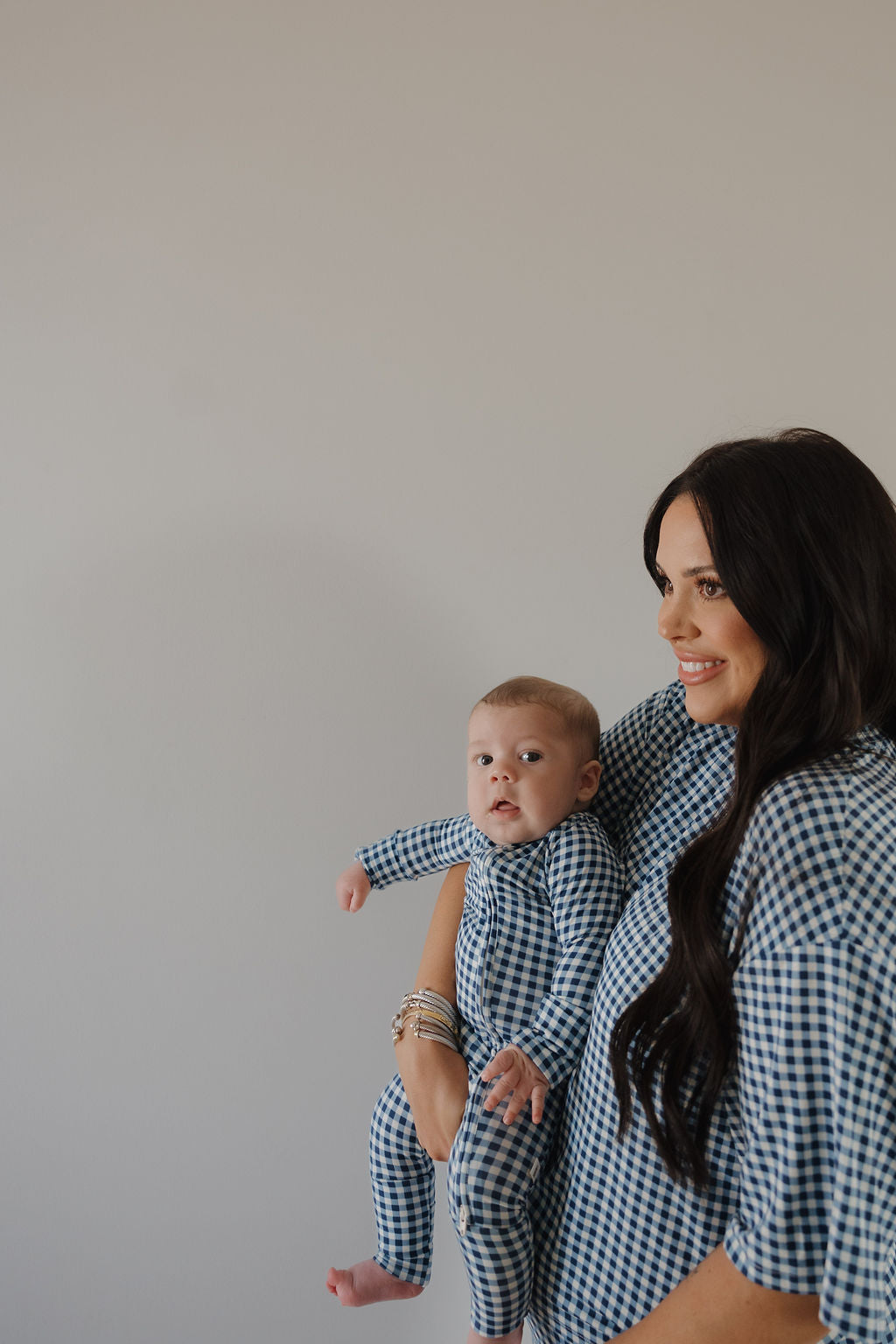 A woman with long dark hair smiles, holding her baby in matching forever french baby's Bamboo Zip Pajamas in Blue Gingham—soft, hypoallergenic sleepwear—against a light background. Both look stylish and comfortable.