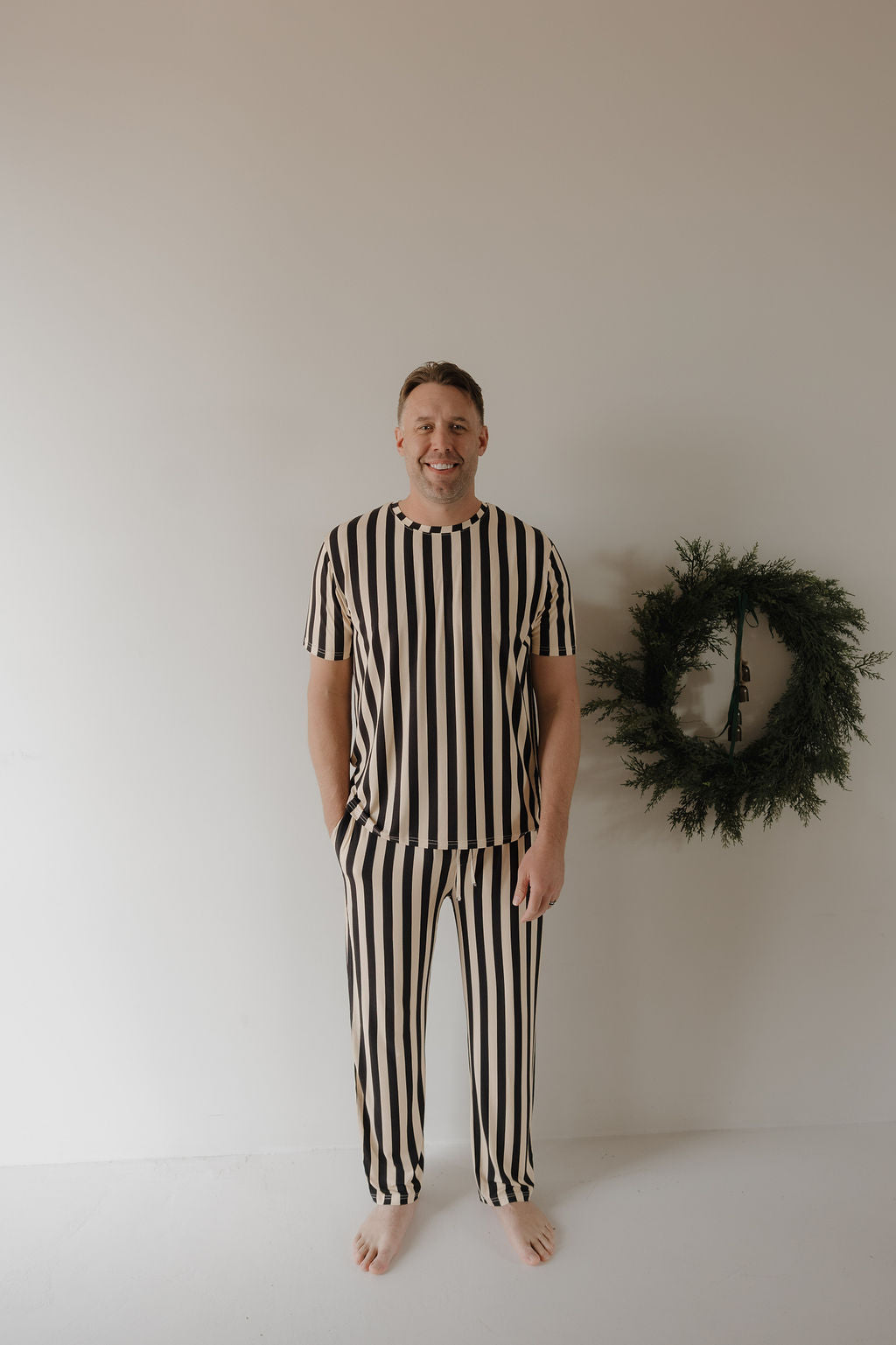 A man smiles barefoot in a minimal room, wearing forever french baby Men's Short Sleeve Pajamas | Midnight Stripe. A simple green wreath on the wall complements his hypoallergenic black-and-white striped pajamas.