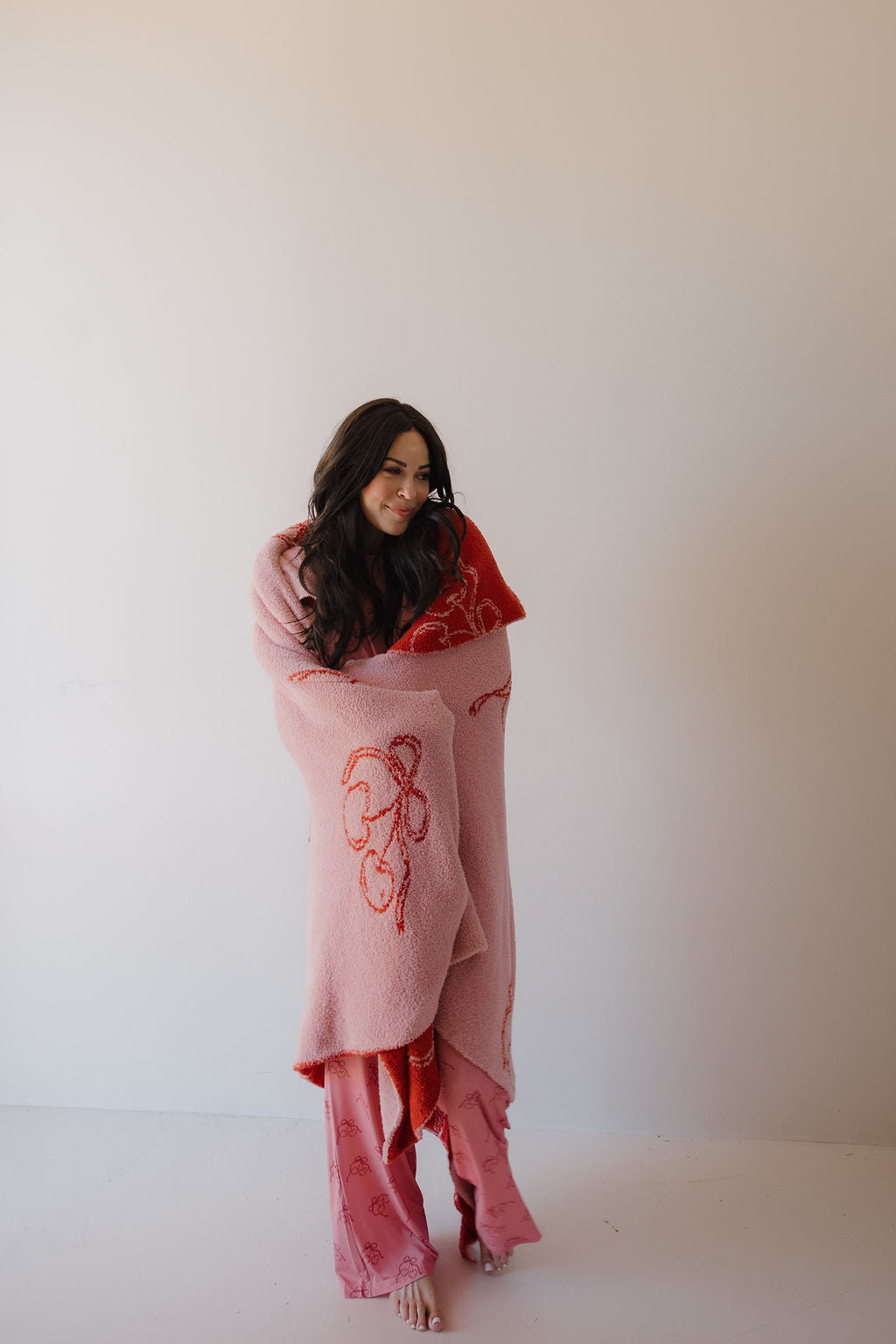 A woman with long dark hair and pink pants stands barefoot against a plain light wall, wrapped in the large Minty Cherries Plush Blanket | Iconic Cherry, featuring Oeko-Tex certified fabric and abstract pink and red patterns.