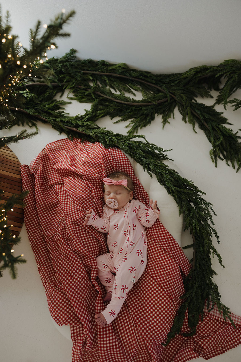 A baby wearing forever french baby Bamboo Zip Pajamas in Twisted Peppermint with a matching headband sleeps on a red gingham blanket, surrounded by pine branches and holiday lights near the Christmas tree.