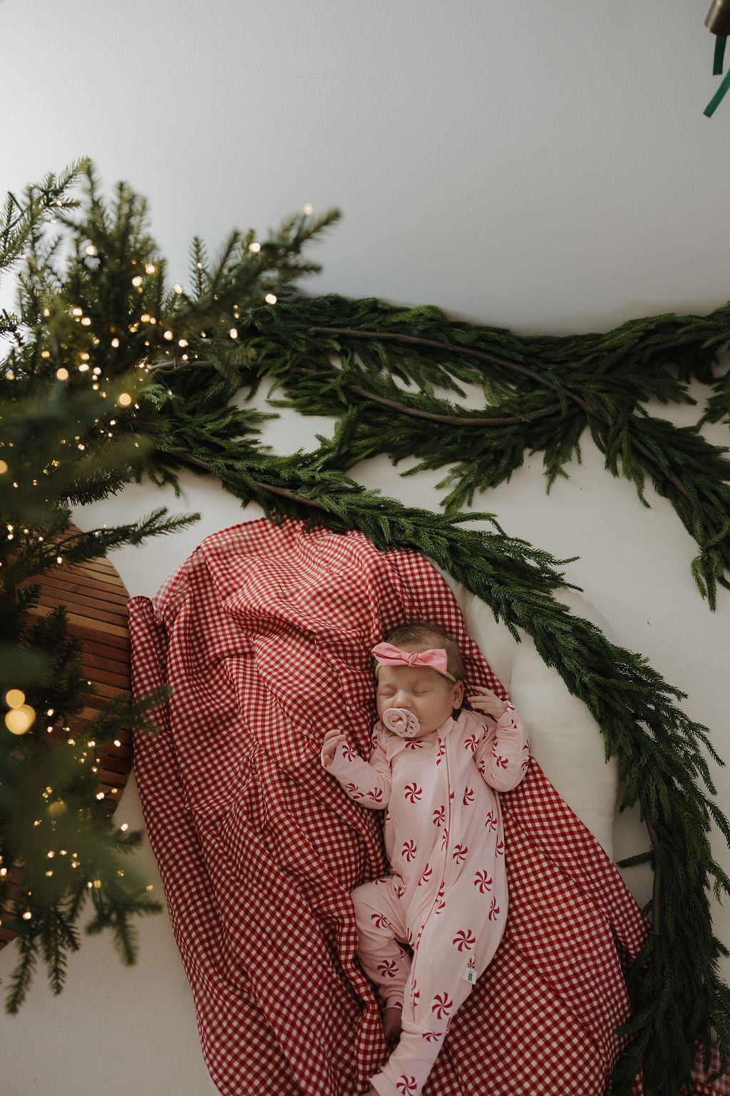 A baby sleeps peacefully on the forever french baby Muslin Swaddle in Red Gingham, wearing a white onesie with red peppermint patterns and a pink bow headband, surrounded by Christmas greenery and twinkling lights.