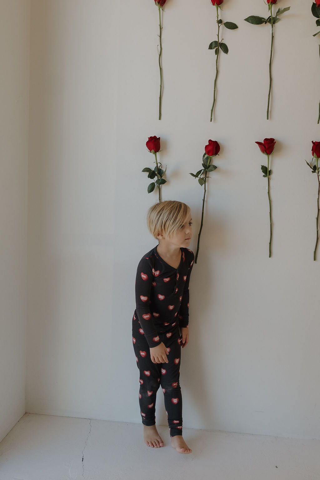 A young child in forever french baby Bamboo Two Piece Pajamas | Love Notes – Charcoal stands barefoot against a white wall adorned with vertical rows of red roses.