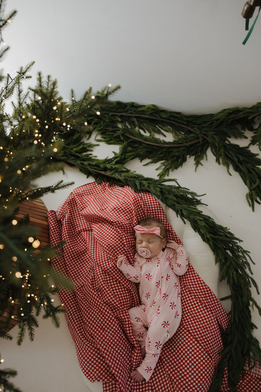 A baby in forever french baby's Muslin Swaddle | Red Gingham naps on a festive blanket next to holiday greenery and a glowing Christmas tree, highlighting cozy, seasonal charm.