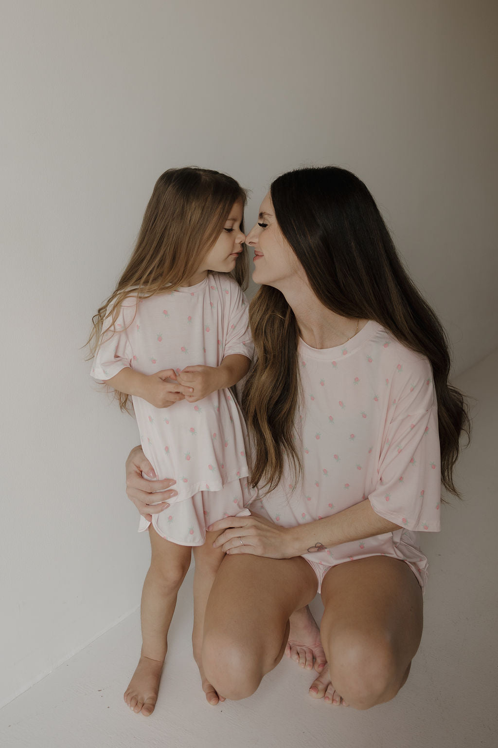 A young girl with long brown hair, dressed in the Girl’s Bamboo Short Set | Rosie by forever french baby, kneels close to a woman as they touch noses and smile softly in a minimalist, light-colored room.