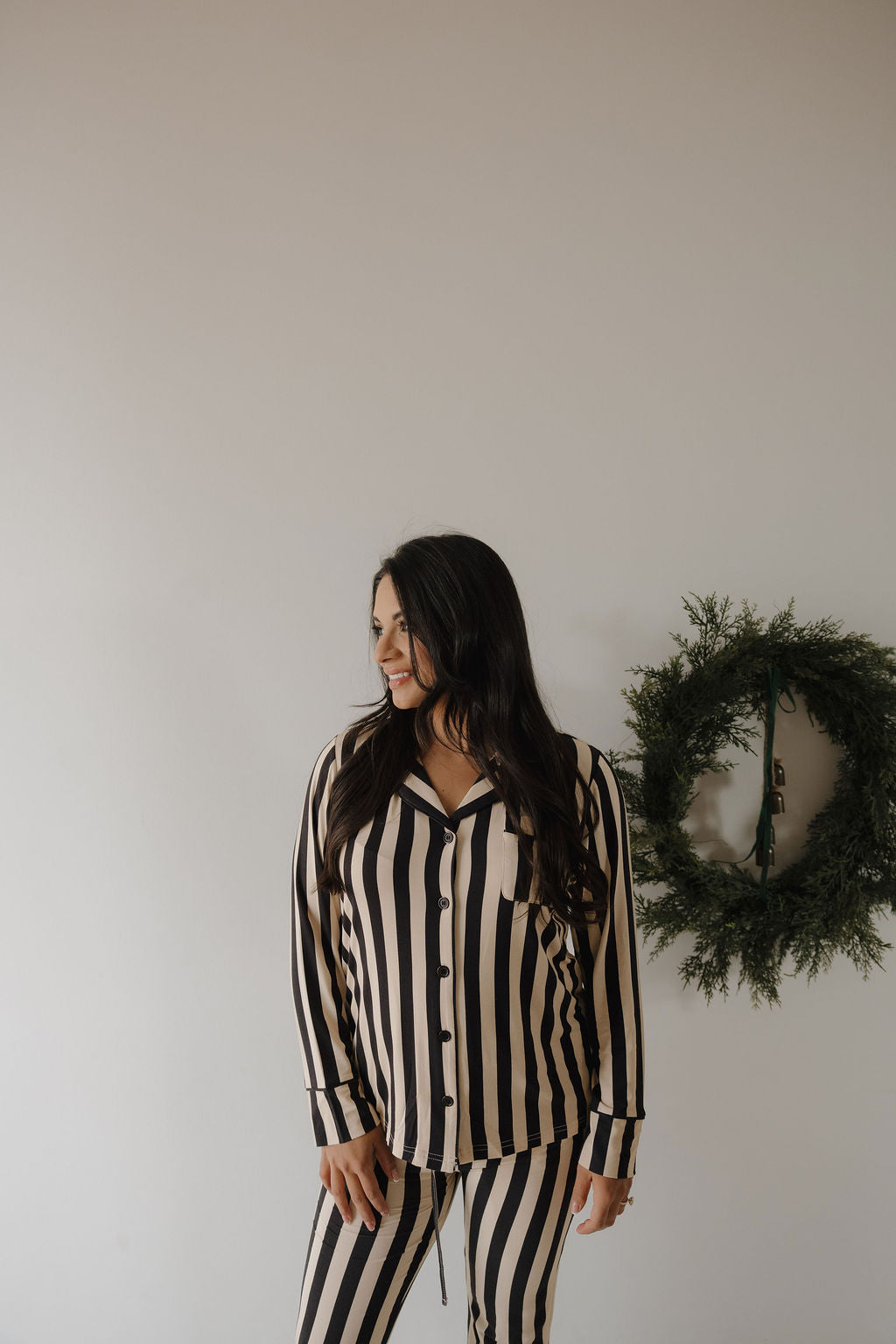 A woman with long dark hair smiles indoors, wearing the forever french baby Women's Bamboo Button Up Lounge Set in Midnight Stripe. The hypo-allergenic outfit is accented by a green wreath on the plain white wall behind her.