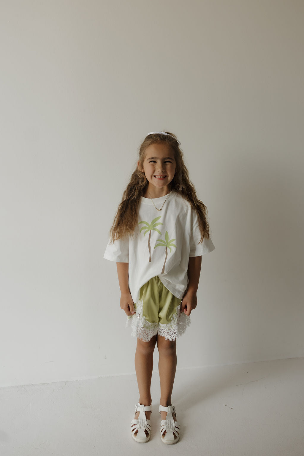 A young girl with long hair stands smiling against a plain white wall, wearing the forever french baby Child Cotton Tee | Palm—crafted from 100% cotton and adorned with palm trees—paired with light green lace-trim shorts and white sandals, making it a perfect choice for coordinated family outfits on sunny days.