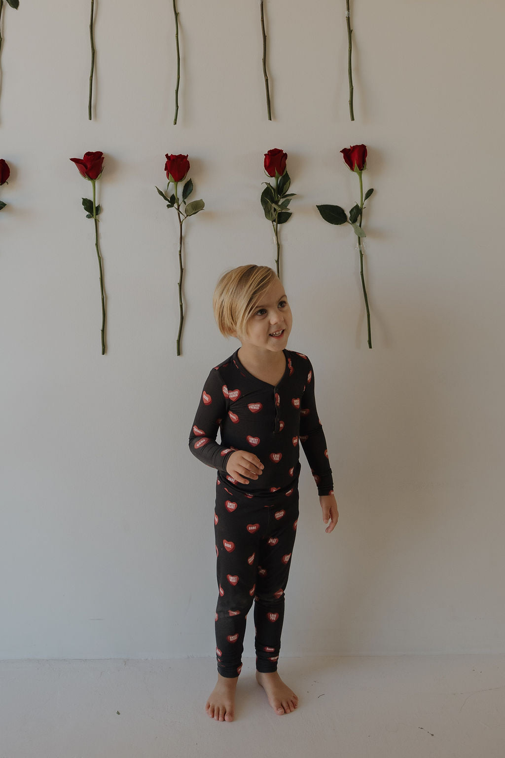 A young blonde child in forever french baby Bamboo Two Piece Pajamas | Love Notes – Charcoal stands barefoot by a white wall adorned with evenly spaced vertical rows of single red roses.